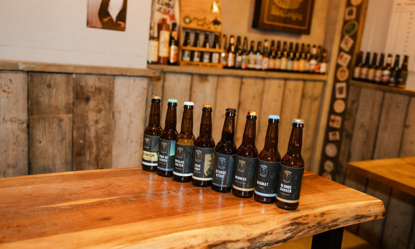 Acht Hong-bierflesjes op een houten tafel in een warme, rustieke proefruimte.