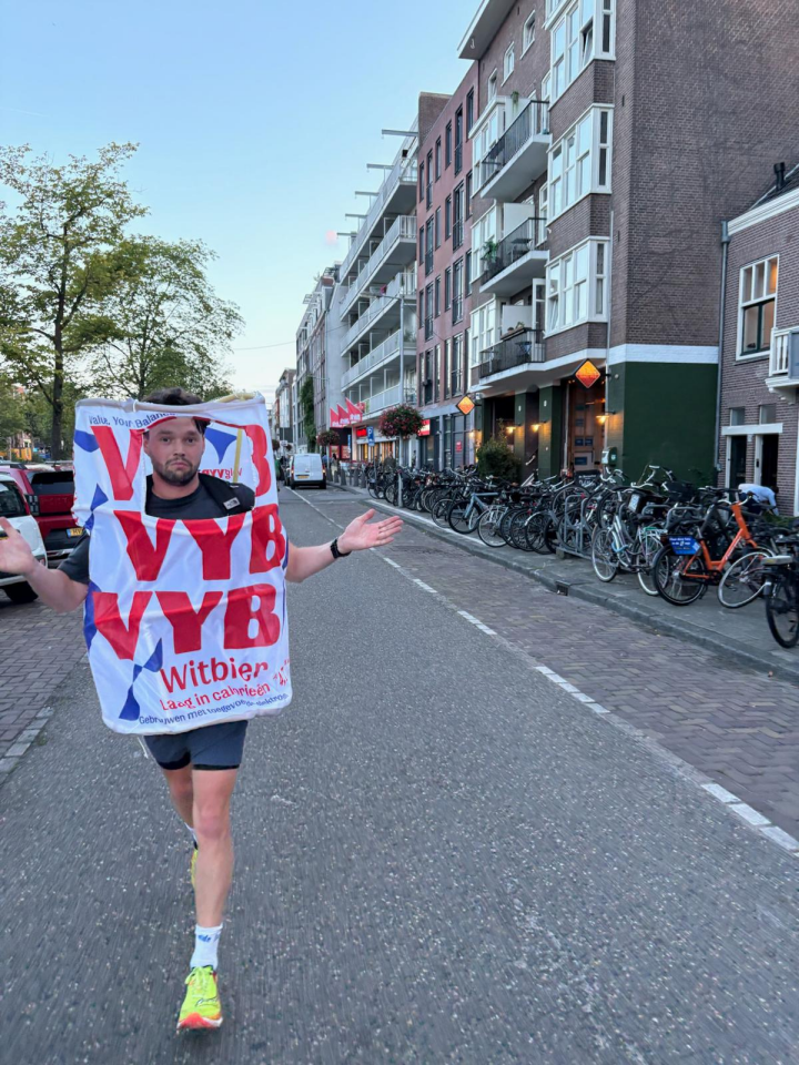 Persoon loopt op straat in kostuum bedrukt met 'VYB' en 'Witbier', fietsen en gebouwgevels op de achtergrond
