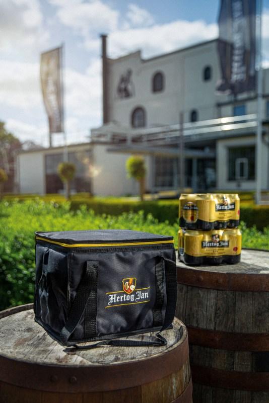 Hertog Jan tas met op de achtergrond blikjes bier en de Hertog Jan brouwerij