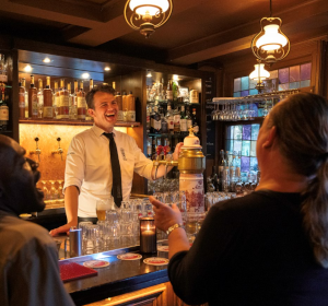 Barman en stamgasten lachen om een grap van een stamgast aan de bar in een Amstel café.