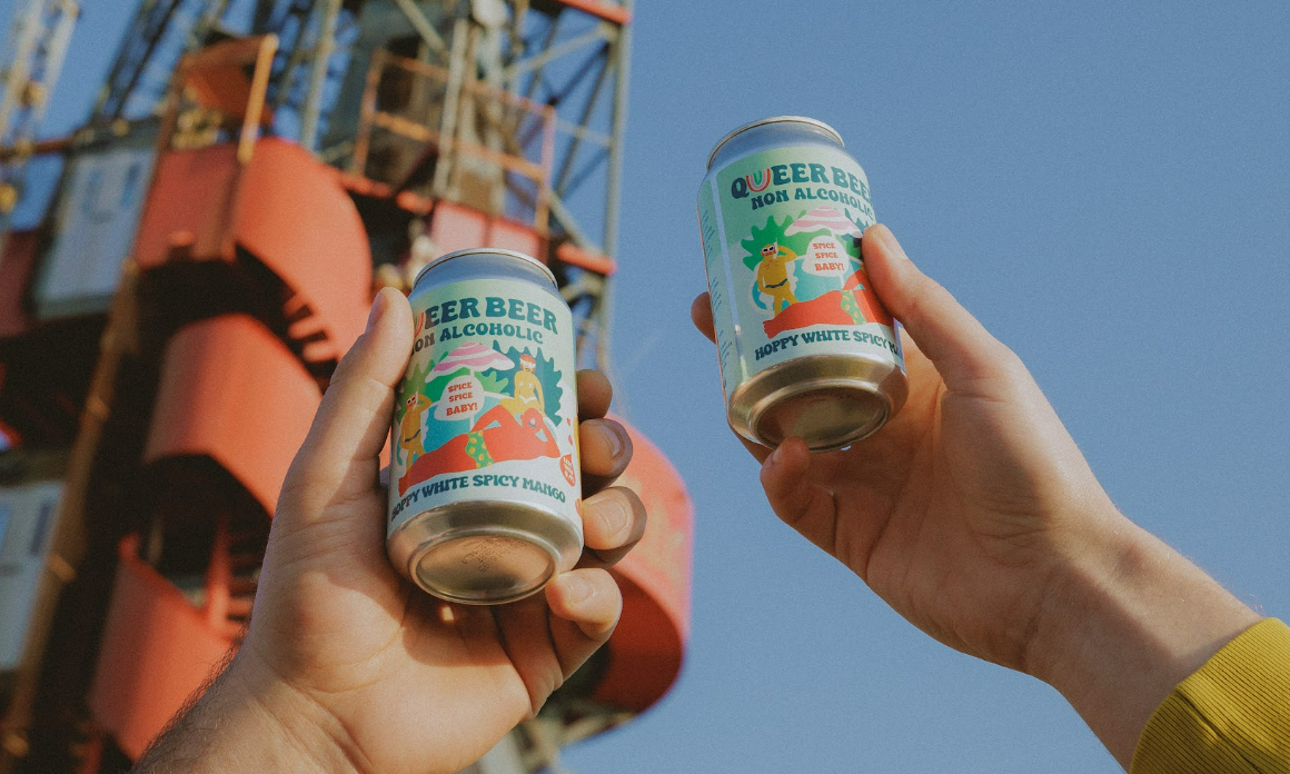 Twee handen houden Queer Beer 0.0 blikken omhoog, met een industriële achtergrond.