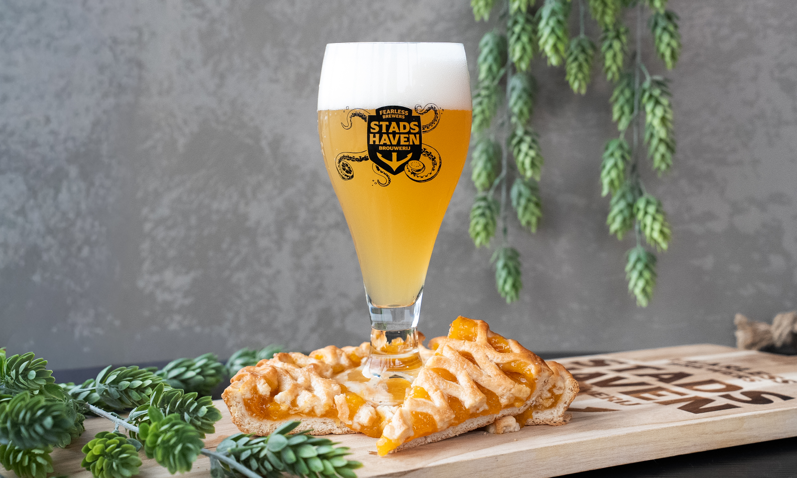 Bierglas met schuim en logo, omringd door groene bladeren en een stuk appelgebak.