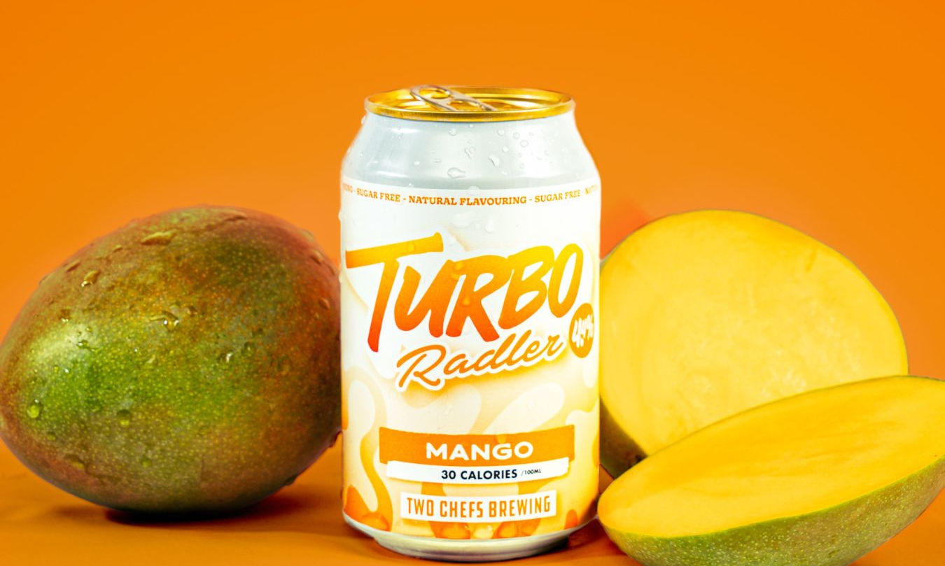 Blikje Two Chefs Turbo Radler Mango tussen twee mango's op een oranje achtergrond.