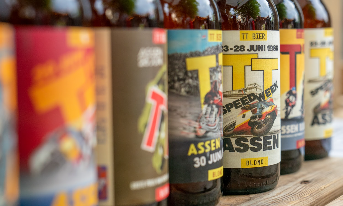 Bierflesjes met kleurrijke TT Assen etiketten op een houten tafel.