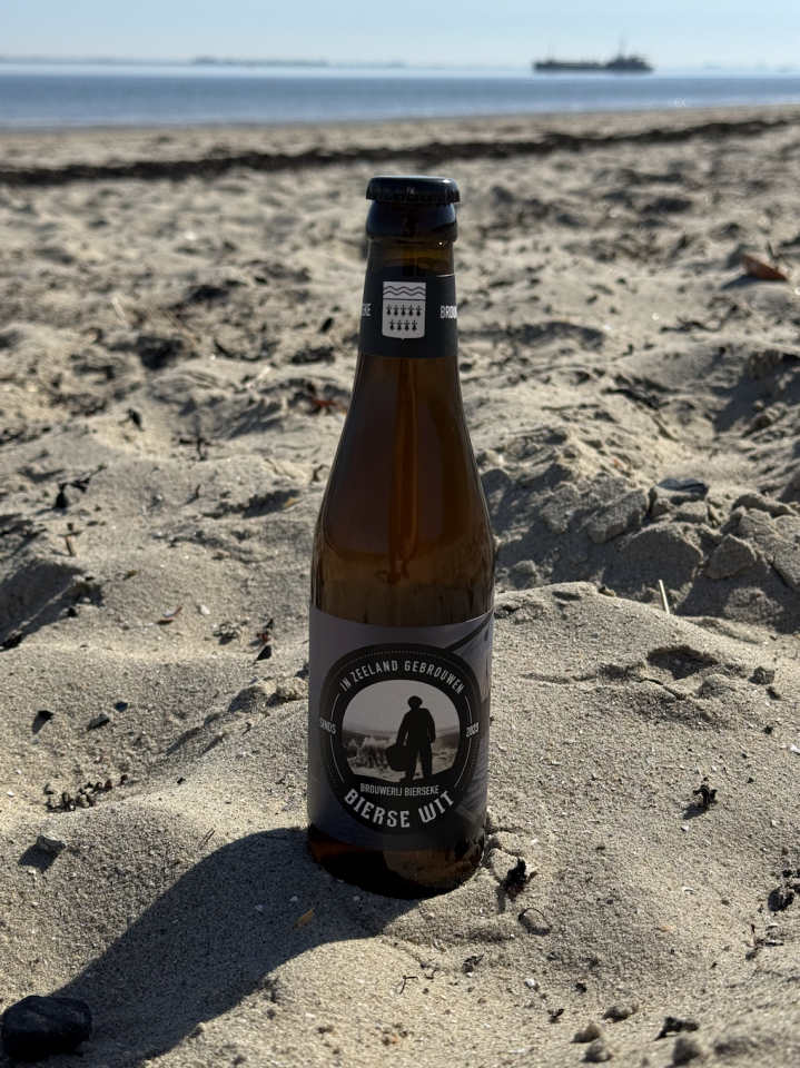 Bierse Wit flesje gedeeltelijk in het zand op een strand met de zee op de achtergrond.