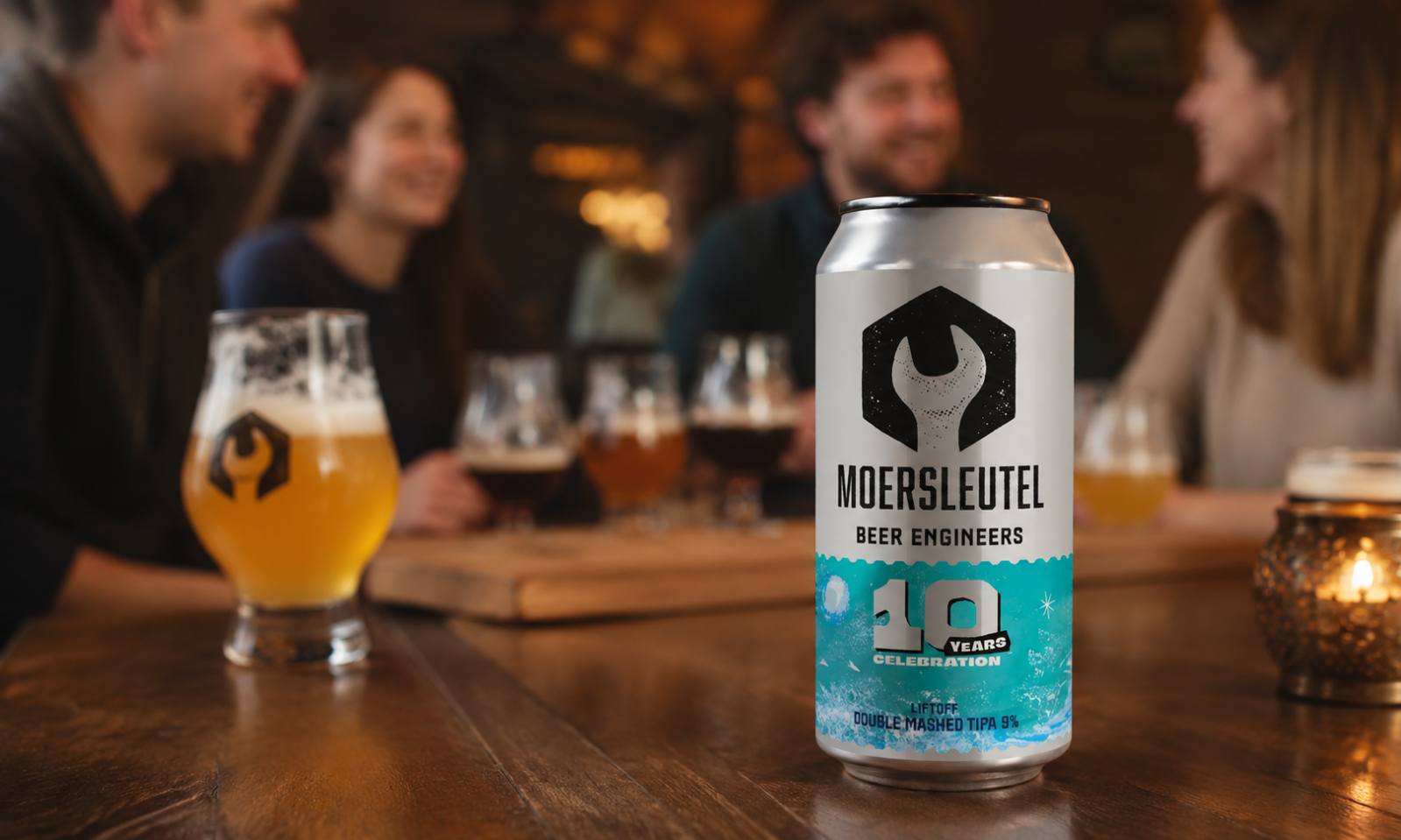 Blik Moersleutel 10 Years Liftoff op houten tafel met bierglazen en onscherp cafégezelschap.