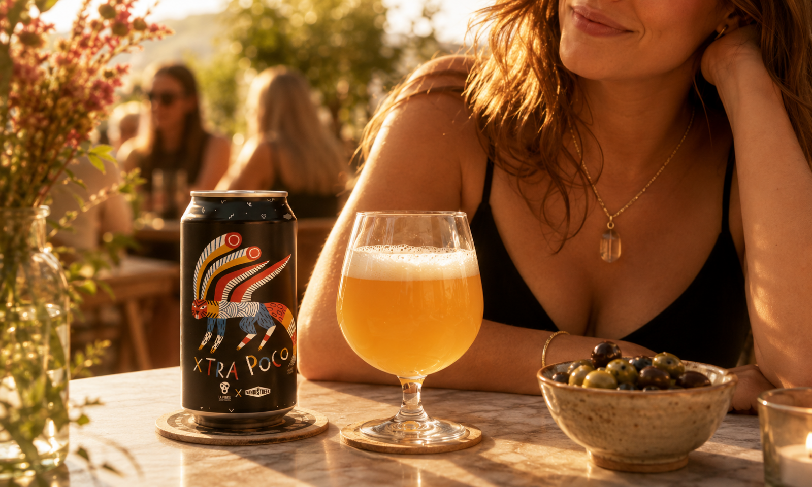 Blik vandeStreek Xtra Poco naast glas troebel bier op zonnig terras met vrouw en olijven.
