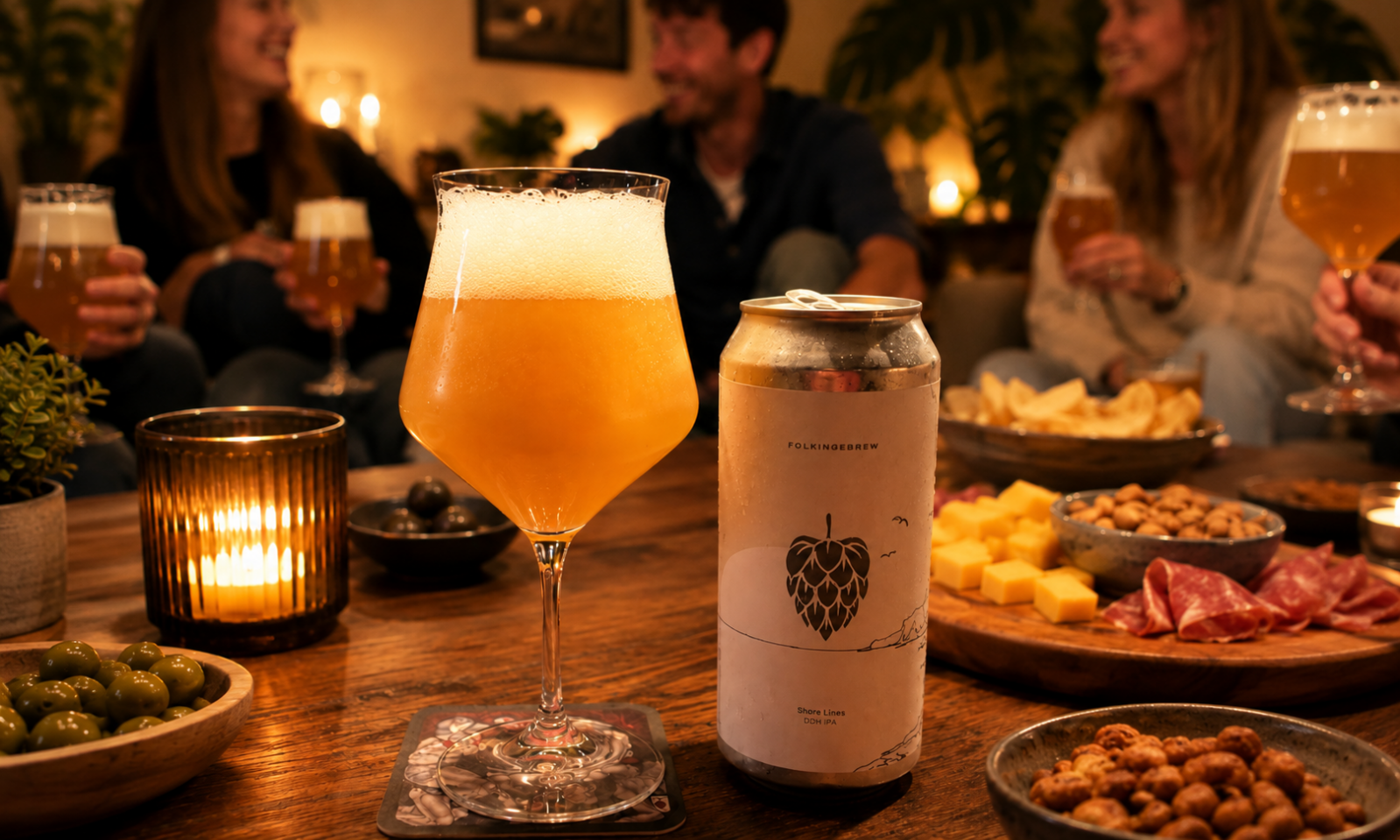 Glas hazy IPA en blik Folkingebrew Shore Lines op borreltafel met kaarslicht, snacks en vrienden op achtergrond.