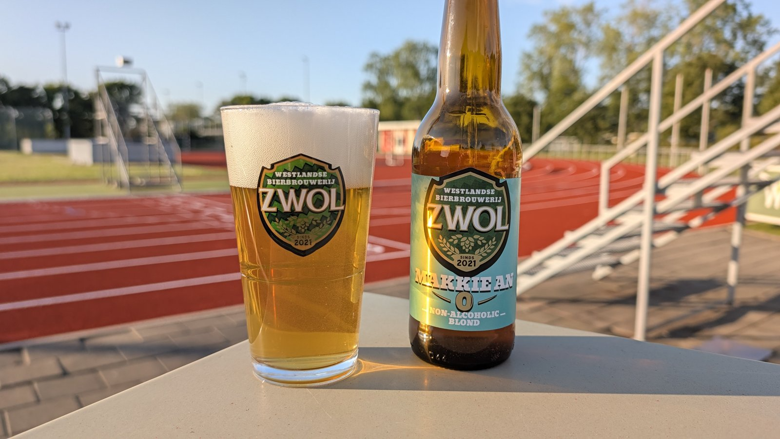 Glas en fles Makkie An alcoholvrij blond bier op een tafel bij een atletiekbaan in de zon.