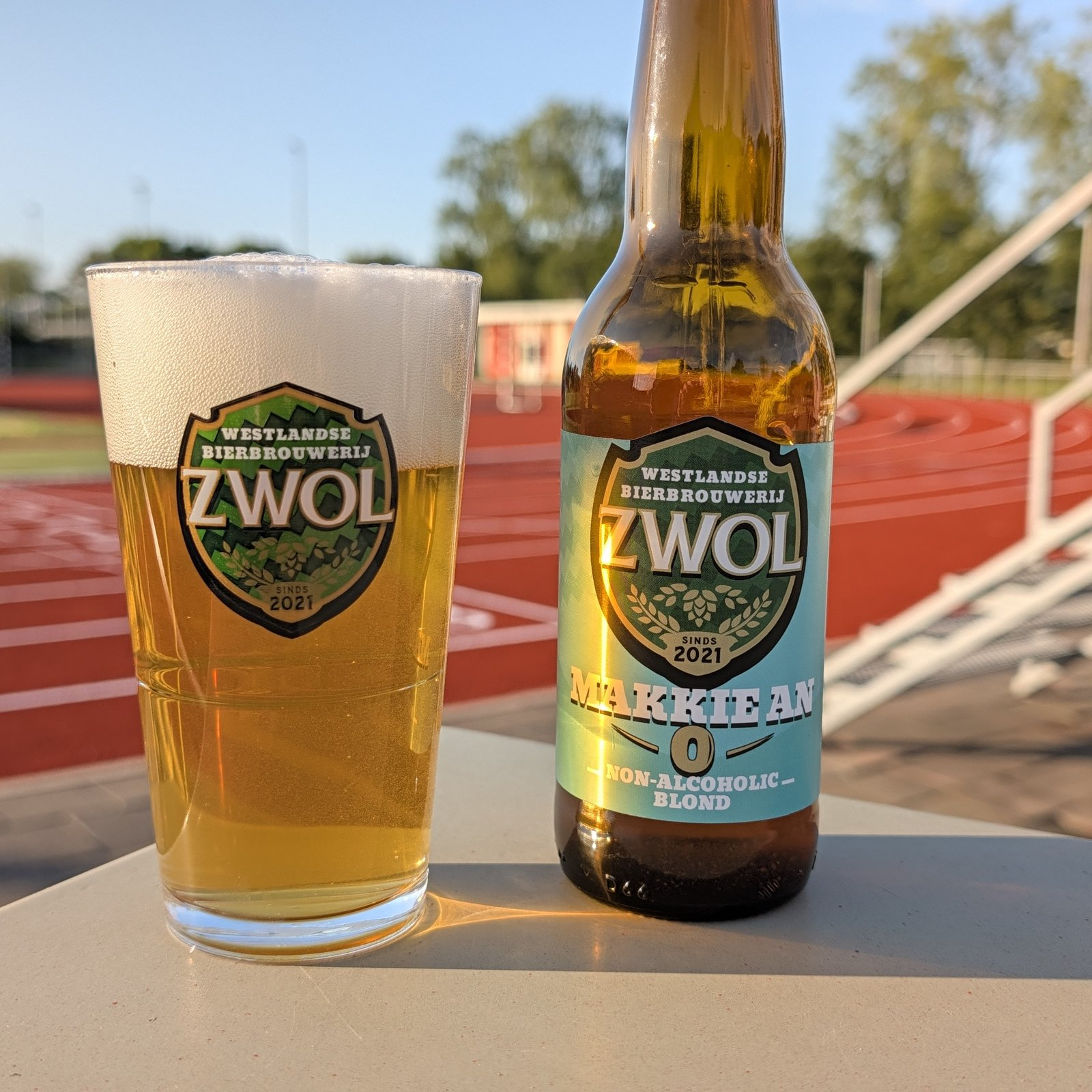 Glas en fles Makkie An alcoholvrij blond bier van Zwol op een tafel bij een atletiekbaan.