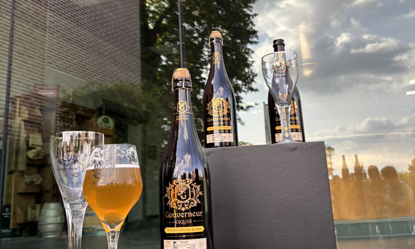Flessen Gouverneur Exquise en bierglazen op zwarte sokkels voor een reflecterende glazen gevel.