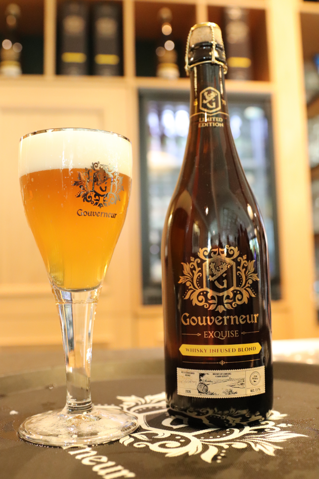 Glas blond bier met schuim naast fles Gouverneur Exquise Whisky Infused Blond op een bar.