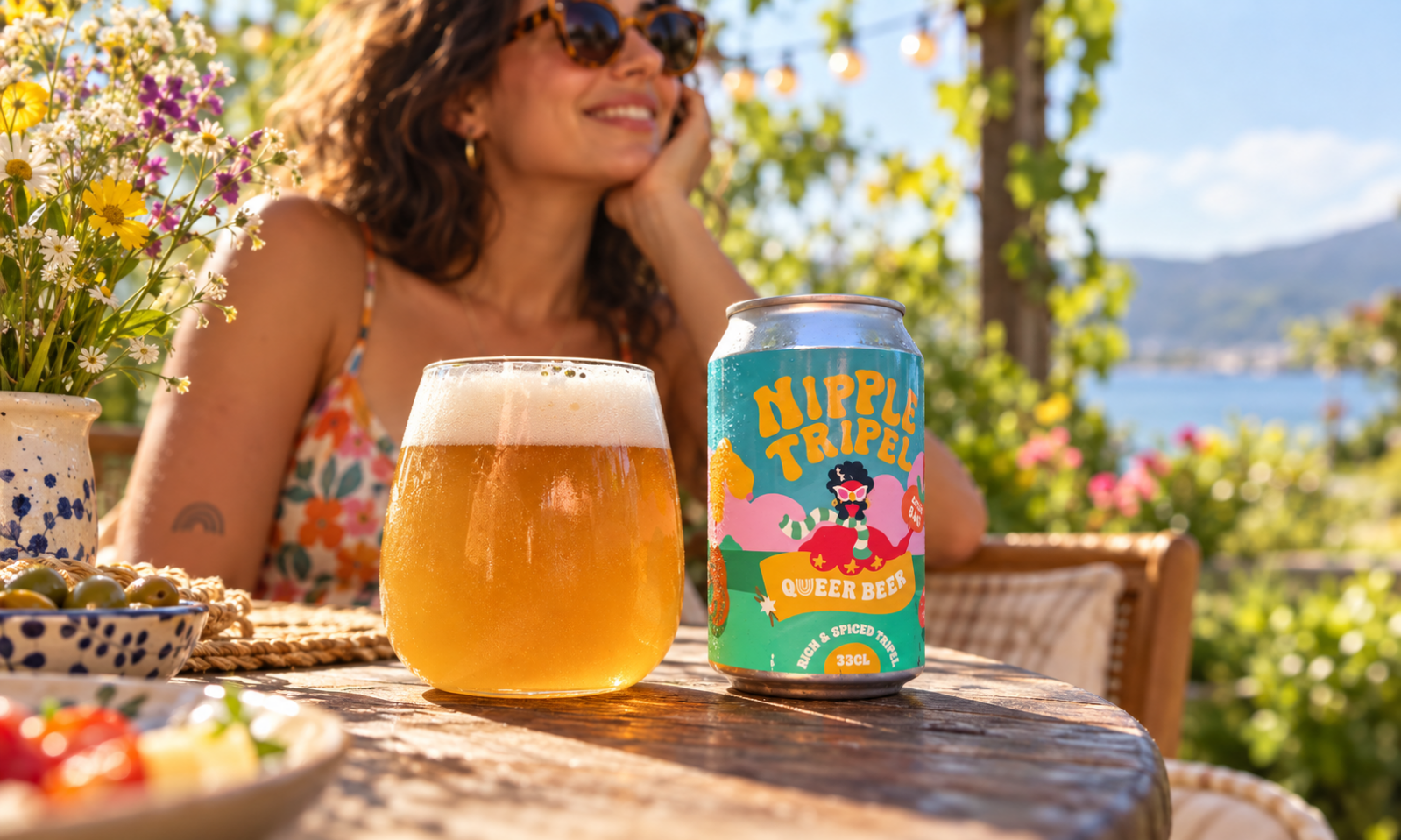 Blik Queer Beer Nipple Tripel met glas blond bier op een zonnig terras aan het water.