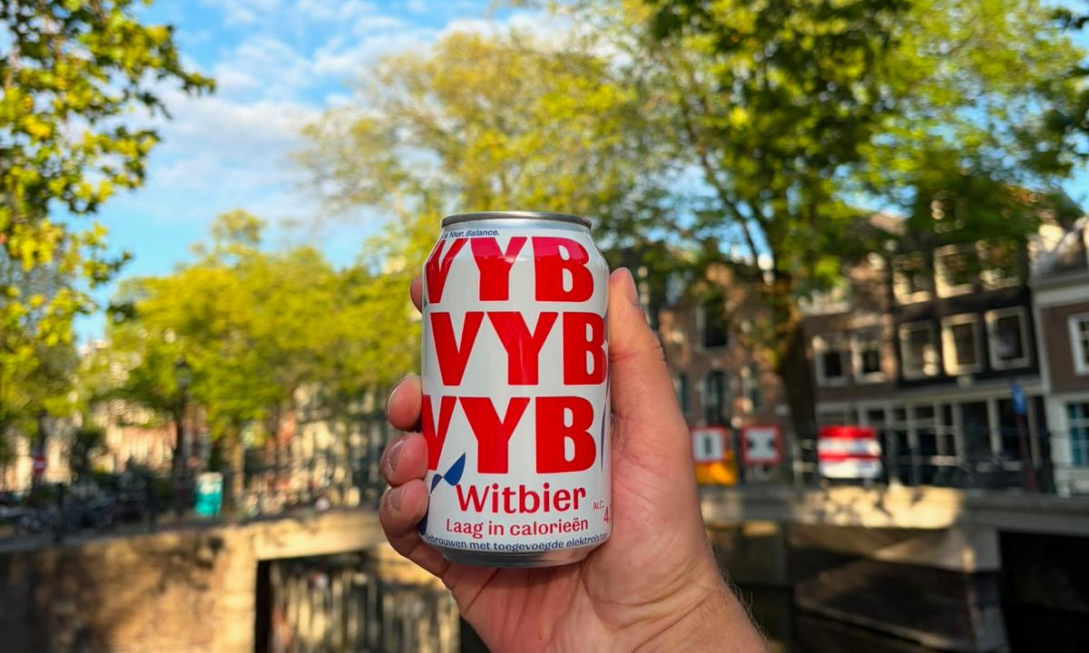 Hand houdt een witte en rode blik VYB Witbier voor een kanaal met bomen en huizen op zonnige dag
