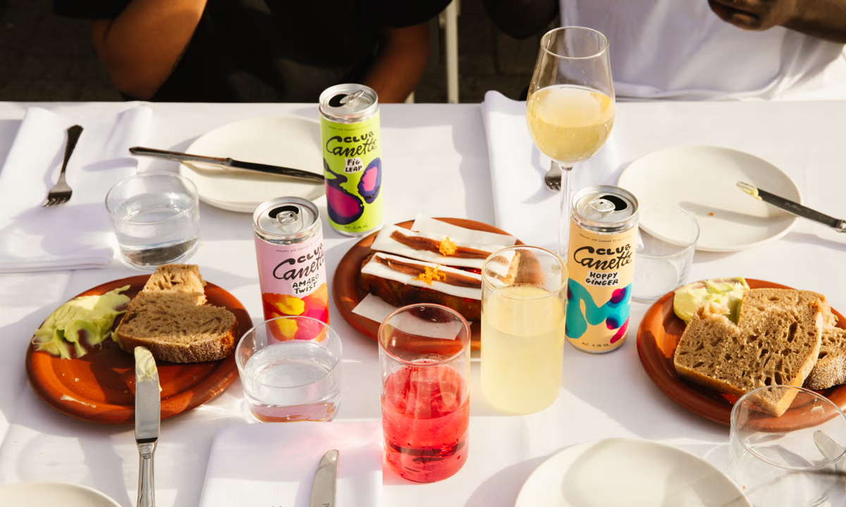 Tafel met drankjes en hapjes, waaronder drie blikjes Club Canette, glazen en borden met brood.