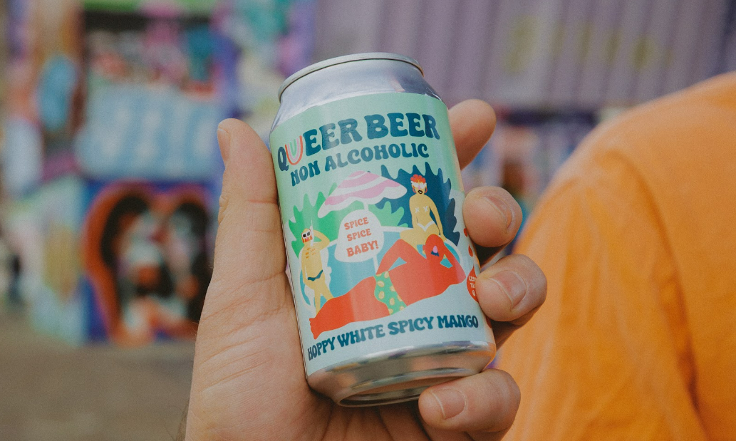 Hand houdt een blikje Queer Beer alcoholvrij bier vast met kleurrijke achtergrond.