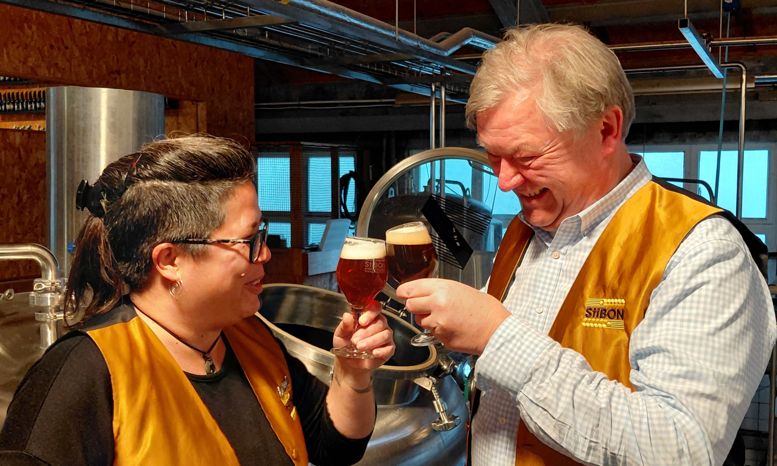 Yu Mey Kwee en Henri Reuchlin proosten met bier in een brouwerijsetting met StiBON-logo op hun kleding.