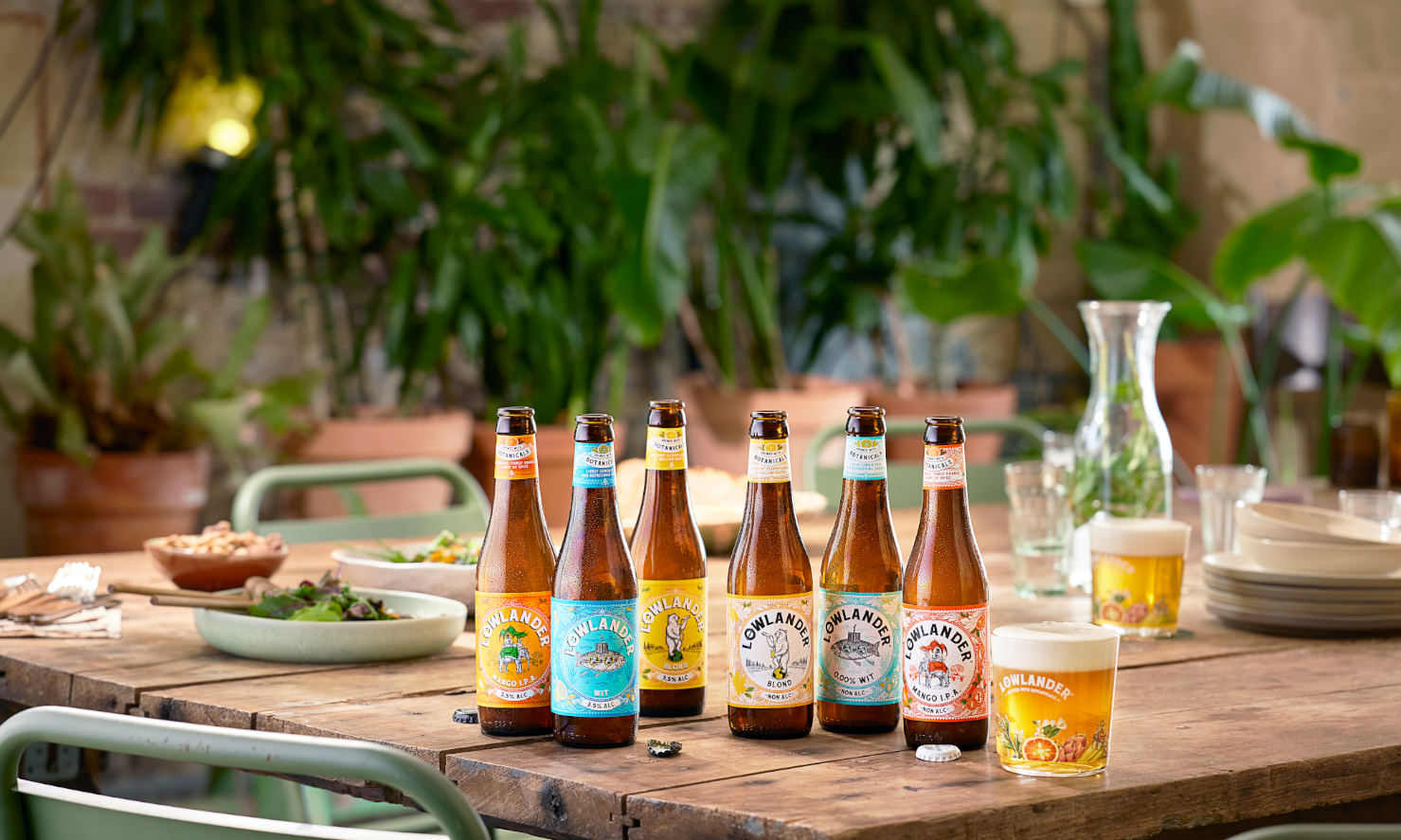 Zes flesjes Lowlander bier op een houten tafel met planten op de achtergrond.