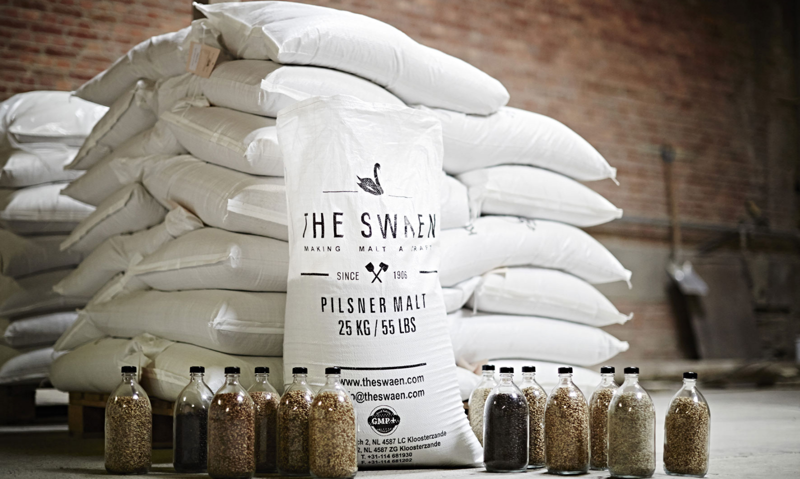 Witte zakken mout met flesjes graan en een bakstenen muur op de achtergrond.