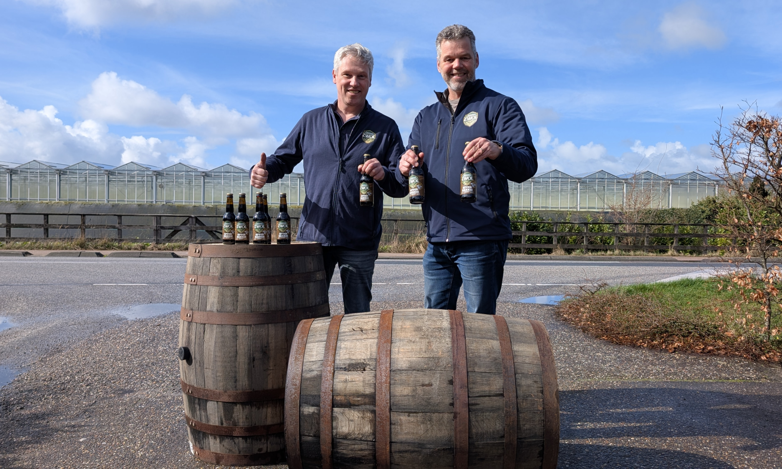 Marty en Ad met Afterbakkie Barrel Aged bierflessen naast houten vaten en kassen op de achtergrond.