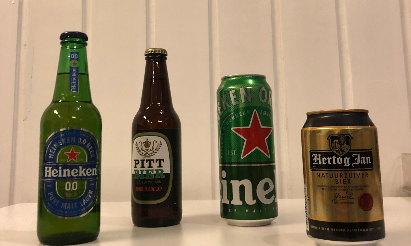 Vier soorten bier op een witte plank, inclusief Heineken, Pitt Bier en Hertog Jan.