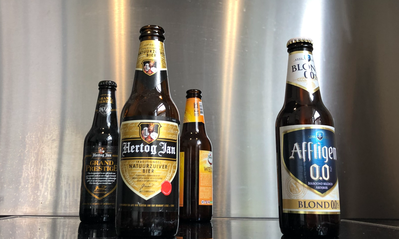 Verschillende bierflessen waaronder Hertog Jan en Affligem op een donkere ondergrond voor een zilveren achtergrond.