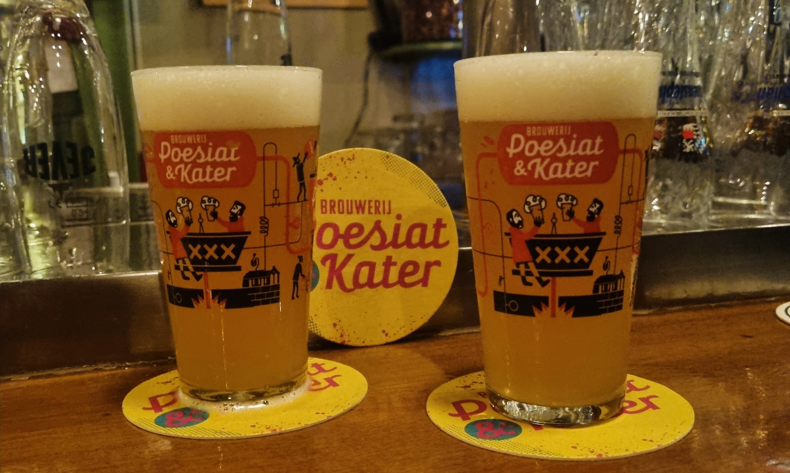 Twee glazen met bier en Poesiat & Kater logo's op een houten bar met een bierviltje.