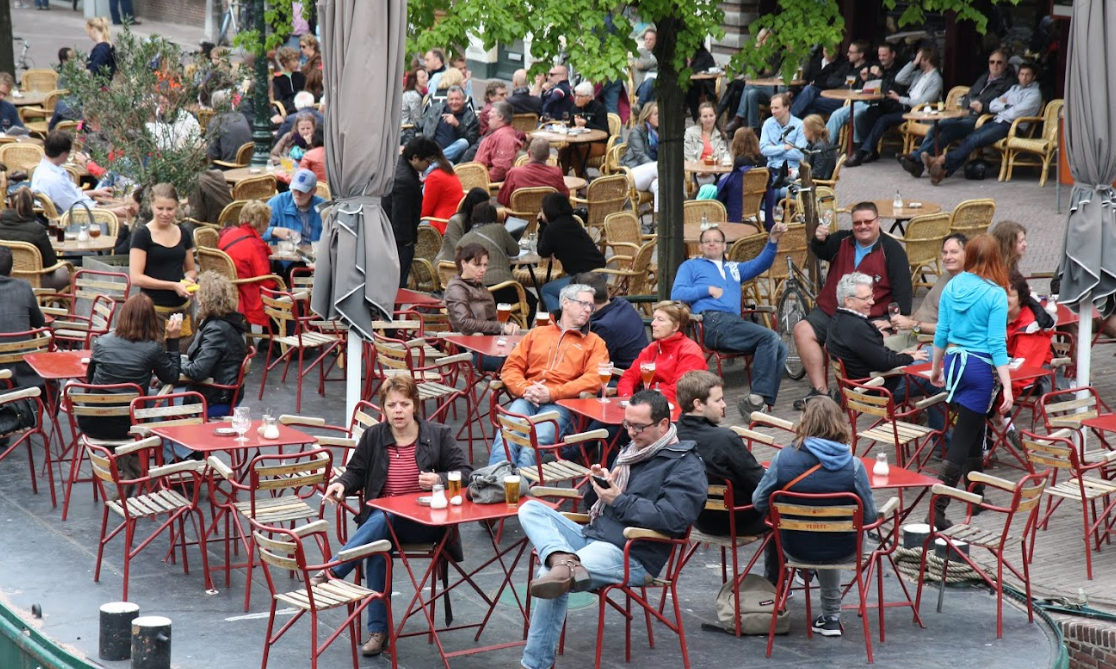 Terrasje vol mensen in de buitenlucht met rode tafels en stoelen.