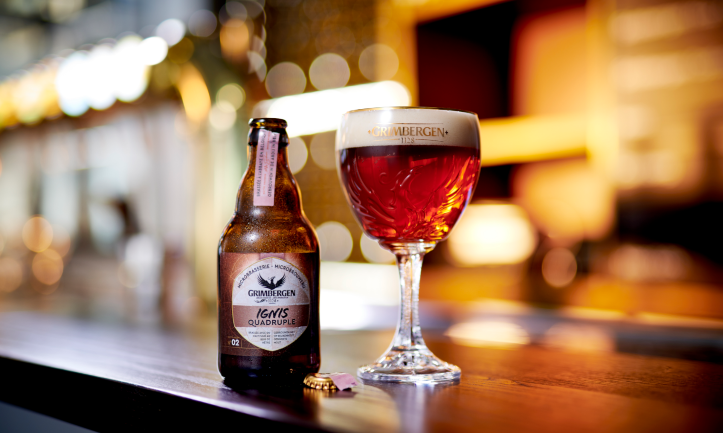 Grimbergen Ignis bierfles en glas bier op een houten bar met onscherpe achtergrondverlichting.