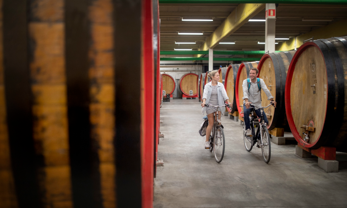 Twee mensen fietsen in Brouwerij 3 fonteinen langs grote houten biervaten (foeders).