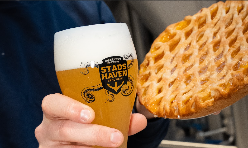 Een hand houdt een glas bier van Stadshaven Brouwerij naast een vlaai vast.