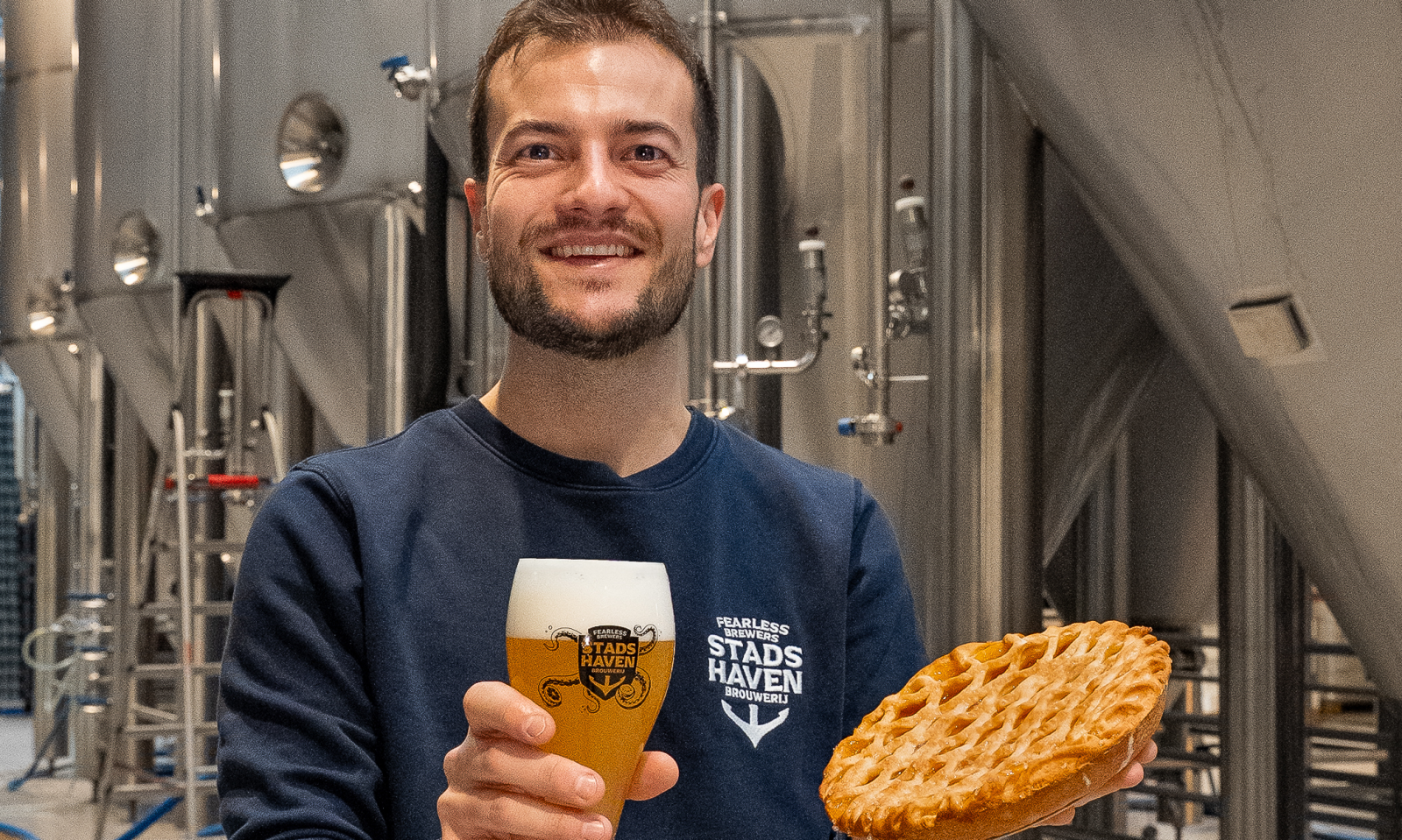 Brouwer Bas Jacob met glas bier en taart in een brouwerij met roestvrijstalen tanks.