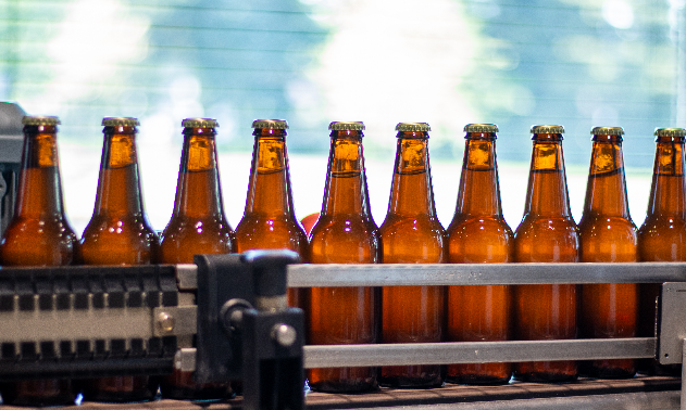Rij van tien amberkleurige bierflesjes op een transportband in een fabriek.