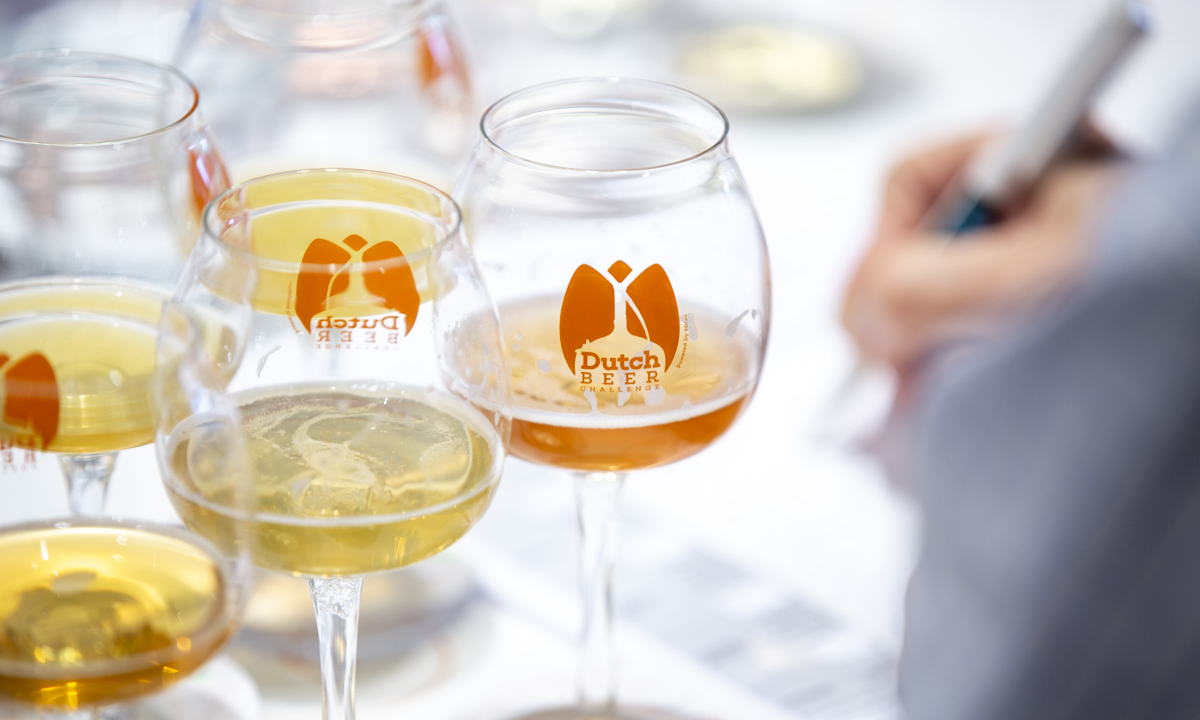 Close-up van bierglazen met 'Dutch Beer Challenge' logo en iemand die aantekeningen maakt.