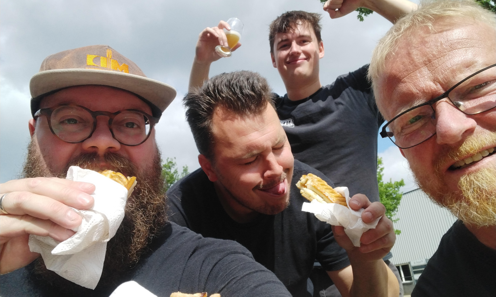 Vier lachende mannen buiten met snacks en bier in een ontspannen sfeer.