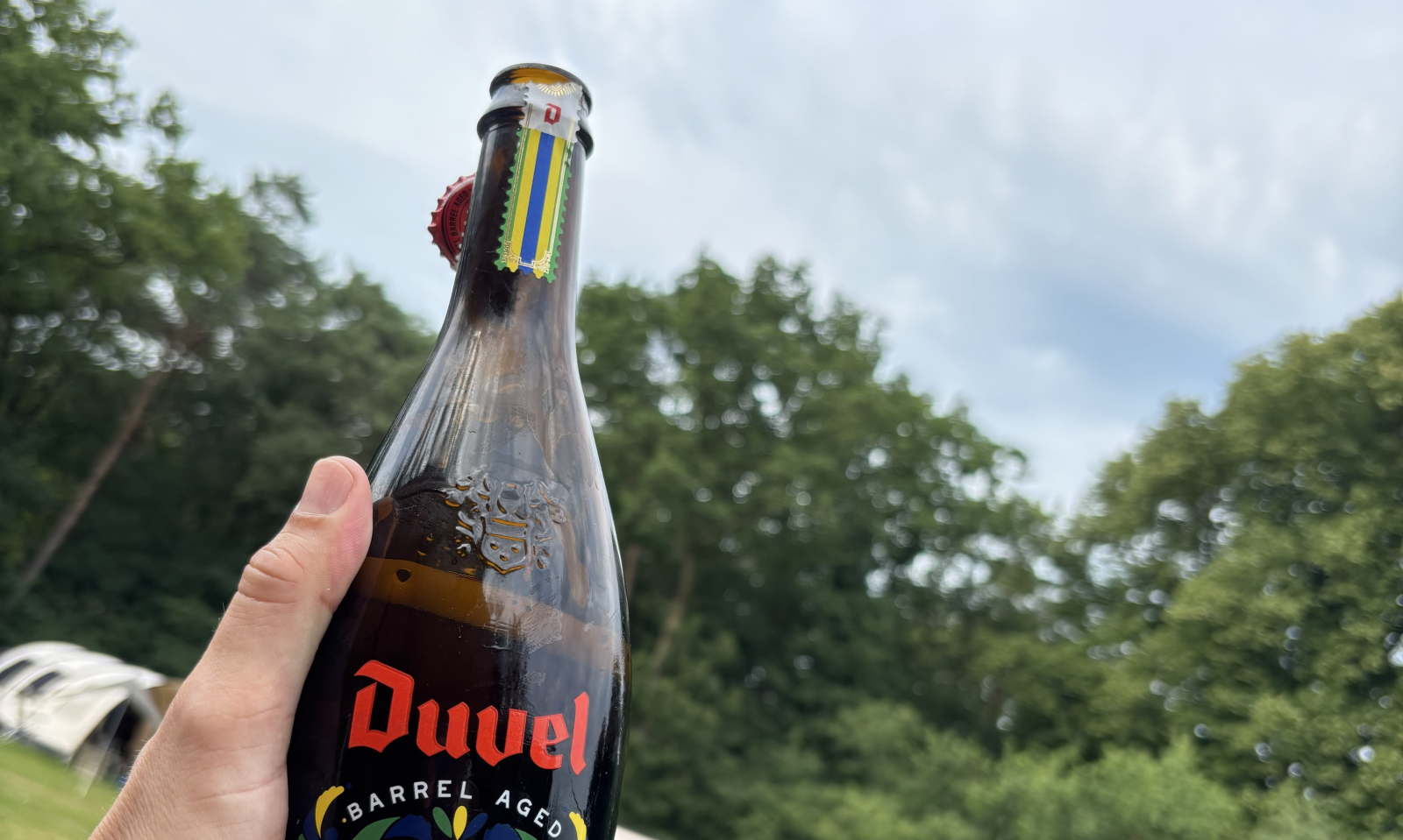 Hand houdt een fles Duvel Barrel Aged Brasil Rhum Edition vast met een kampeerterrein op de achtergrond.