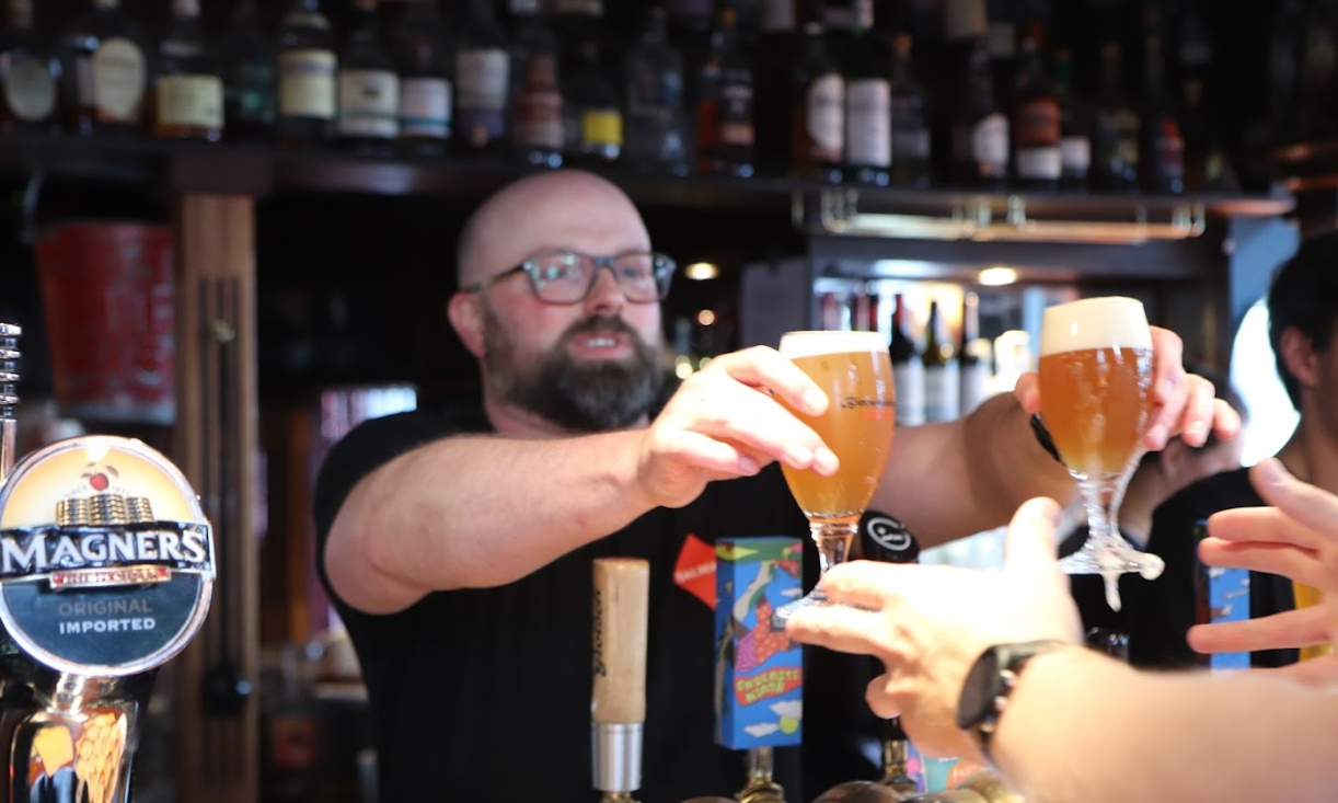 Barman reikt twee glazen bier aan in een drukke kroeg met flessen op de achtergrond.