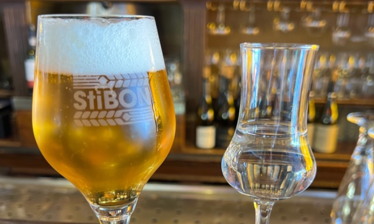Een bierglas en een leeg glas met StiBON logo op een bar met flessen op de achtergrond.