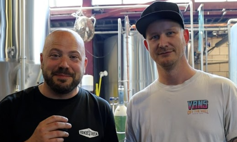 vandestreek en atone brouwers poseren in een brouwerij tussen tanks, met vandeStreek-logo op een zwart shirt.