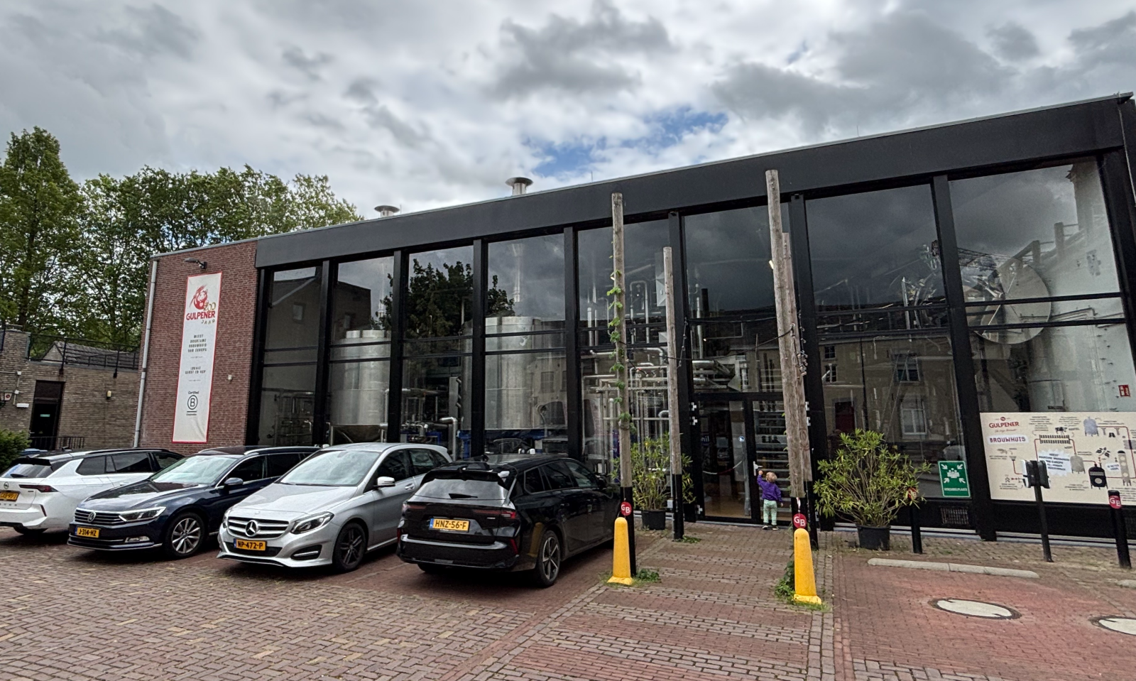 Buitenzijde van de Gulpener-brouwerij met glazen gevel, geparkeerde auto's en bewolkte lucht.