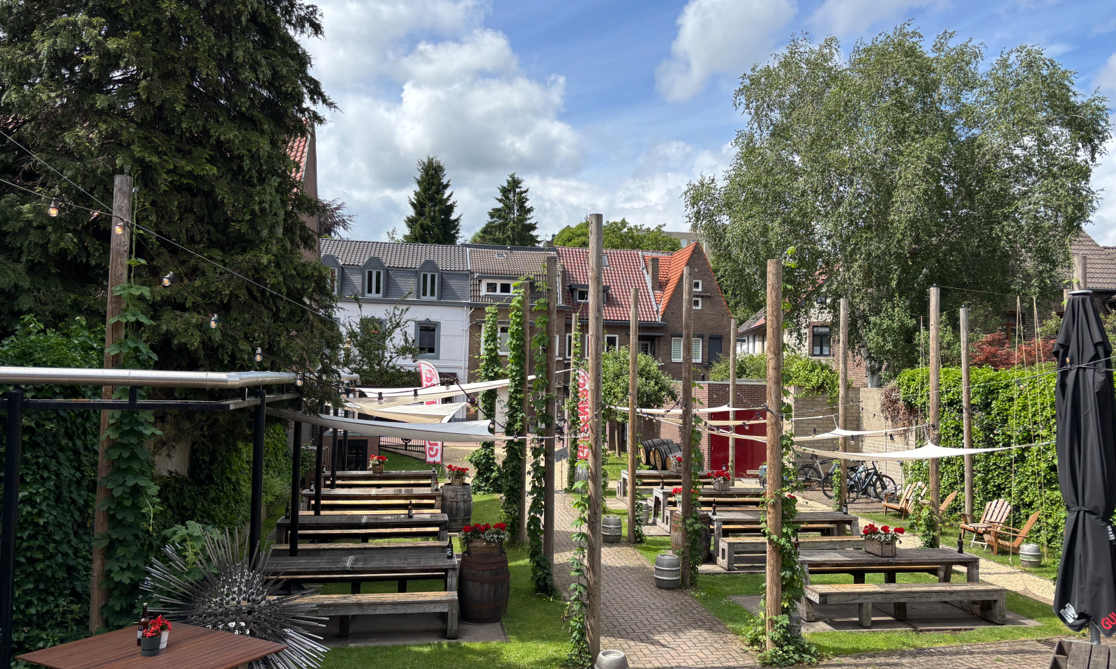 Zonnig bierterras met houten picknicktafels, houten palen, groen en dorpshuizen op de achtergrond.
