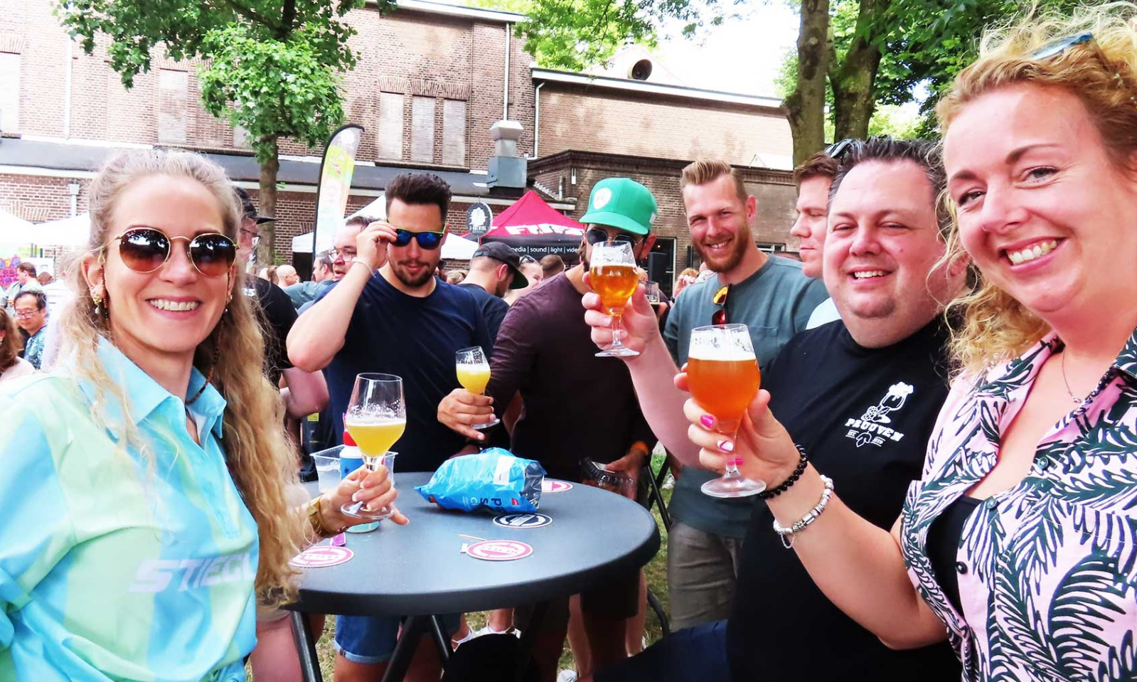 Groep lachende bezoekers proost met bierglazen op een druk buitenfestival in Oss.