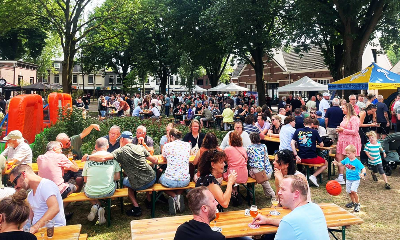 Druk bezocht bierfestival in de buitenlucht met bezoekers aan houten tafels onder bomen en tenten.