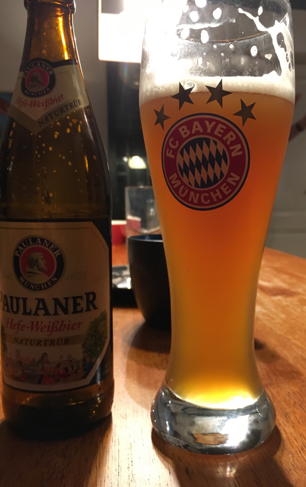 Glas Duits weizen met FC Bayern-logo naast een Paulaner-fles op een houten tafel.