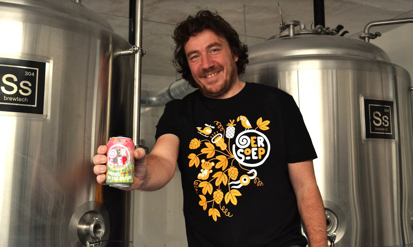 Glimlachende Oersoep-brouwer houdt een bierblik vast tussen roestvrijstalen brouwtanks.