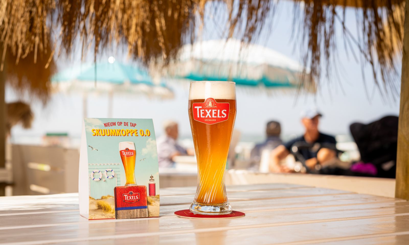 Glas Texels Skuumkoppe 0.0 op een strandterras met promotiekaart en parasols op de achtergrond.