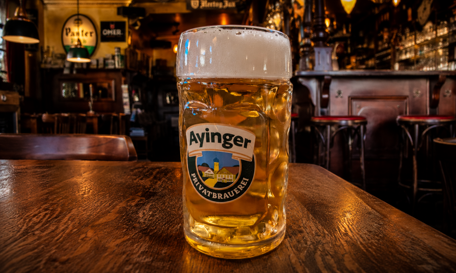 Glas Ayinger bier met schuimkraag op een houten tafel in een warme, traditionele kroeg.