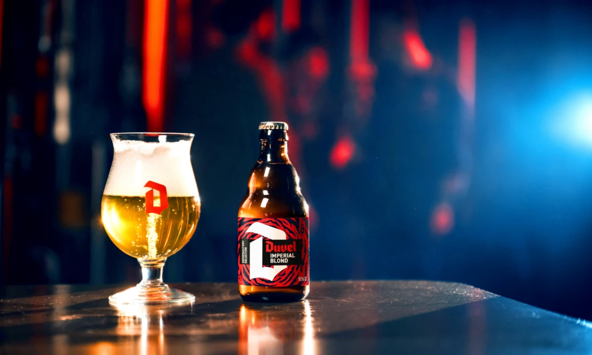 Glas blond Duvel-bier met schuim naast een fles Duvel Imperial Blond op een verlichte bar.