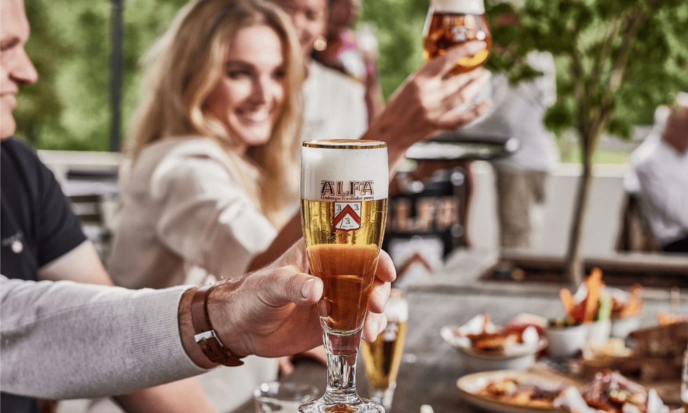 Hand houdt een glas Alfa pils omhoog tijdens een gezellige borrel op een zonnig terras.