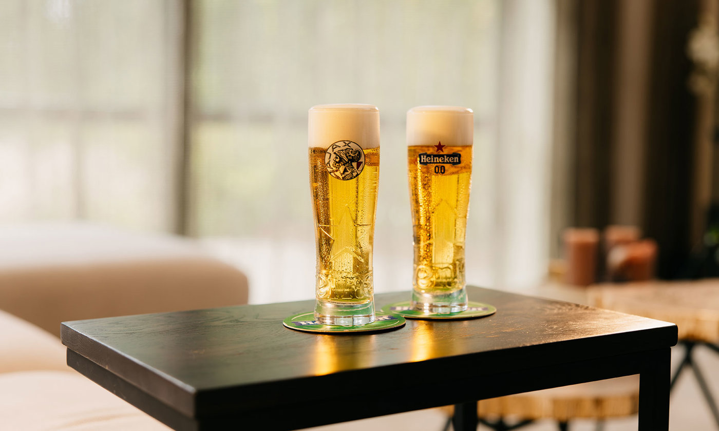 Twee gevulde Heineken 0.0-glazen, waarvan één met Ajax-logo, op een zwarte tafel.